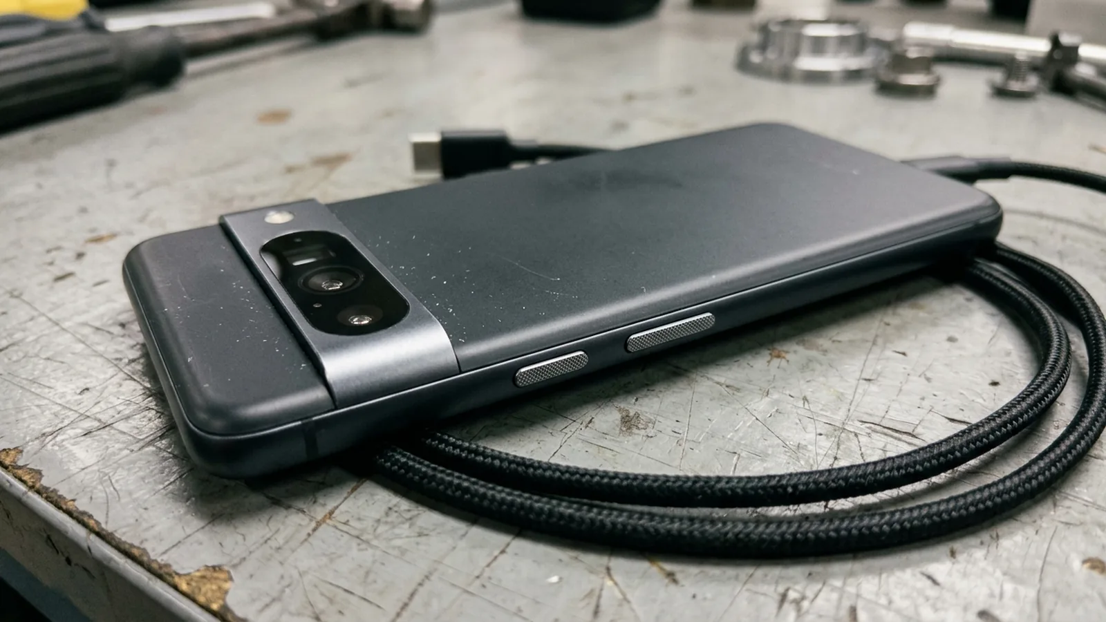 Face-down dark matte Android phone on a grey workbench showing its rear camera bar and side buttons next to a coiled USB-C cable